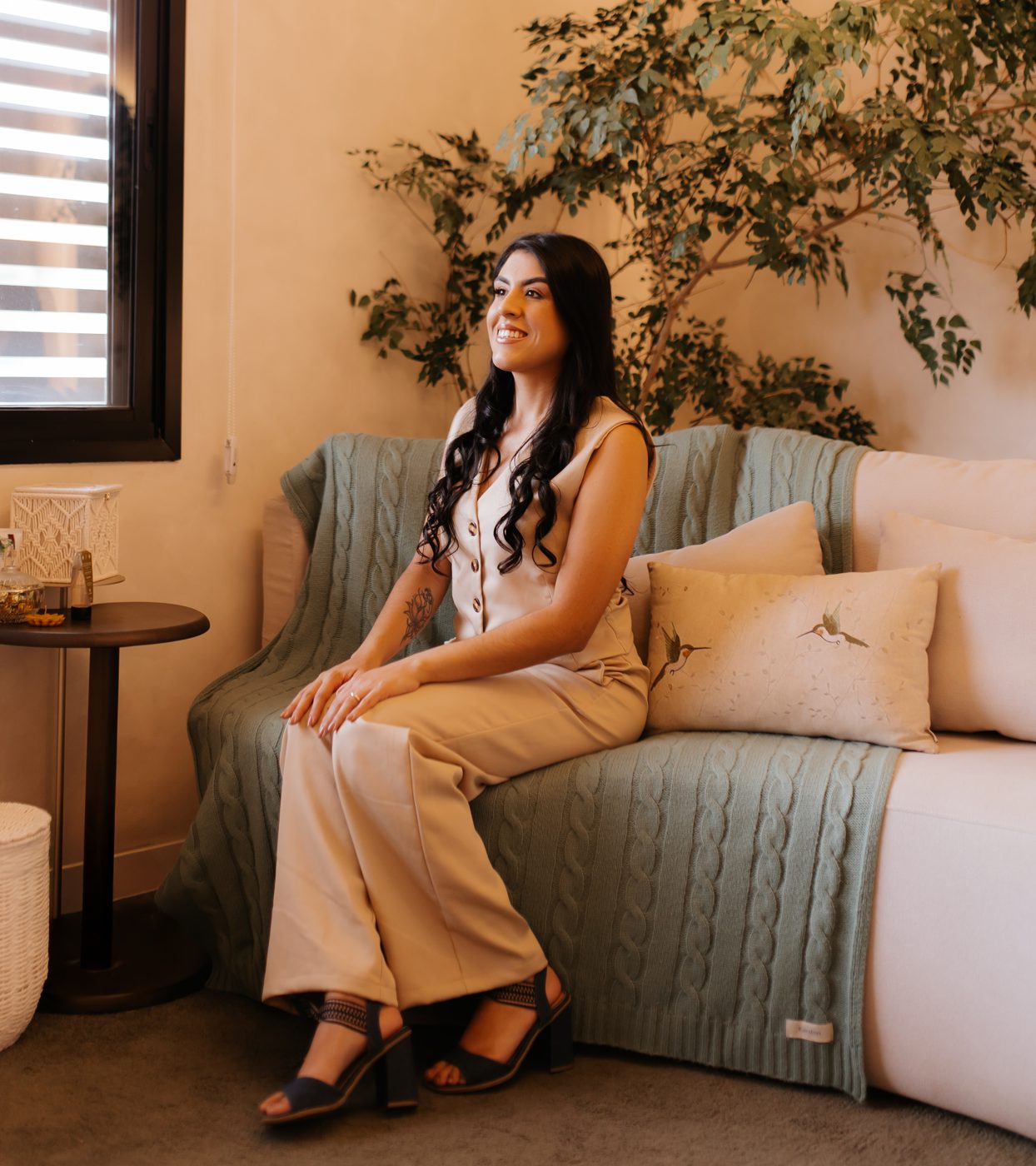 Aline Housni sentada em um sofá, ao lado de uma planta
